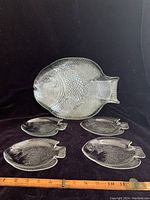 Set of one large fish-shaped glass platter and four smaller fish-shaped glass plates on black background with wooden ruler for scale showing measurements.