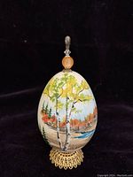 Front view of handpainted egg showing detailed landscape painting with birch trees, water, and autumn scenery, mounted on decorative metal stand and topped with finial.