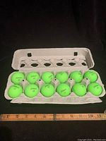 Full dozen of neon green Titleist golf balls neatly placed inside a cardboard container against a black background with a ruler for scale.
