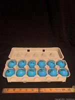 Full view of one dozen blue Nike Mojo golf balls in a beige pulp cardboard egg carton next to a ruler showing scale, each ball marked with Nike Mojo branding.