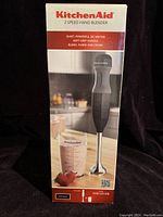 Front view of KitchenAid 2 speed hand blender box showing the blender and included measuring cup with strawberries on the countertop.