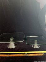 Two clear glass pedestal plates side by side showing size difference with measuring tape on black background.