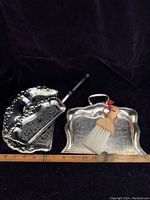 Overall view of two silverplate silent butlers and a small brush with wooden handle, placed on a black background next to a ruler for size reference.