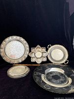 Five vintage silver items laid on black cloth: large round plate with pierced edge, leaf-shaped coaster, round tray with handles, smaller round plate, and round trivet.