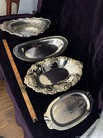 Four vintage silverplate bread trays displayed on a dark cloth background with a ruler for size reference, showing various shapes and decorative styles.