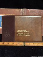 Brown leather-textured Olympic Stamp Souvenir Collection album with gold lettering and embossed Olympic rings, positioned above a ruler for size reference.
