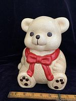 Front view of vintage ceramic teddy bear cookie jar showing face, red bow, and paw details with a measurement scale in front.