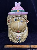 Front view of vintage ceramic cookie jar showing winking monkey face with pink 'COOKIES' hat and flower detail.