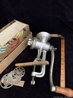 Vintage Perfection 65 food grinder with clamp base, crank handle, extra grinding discs and original colorful box shown together on black background.