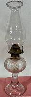 Full view of the oil lamp showing the tall clear glass chimney, ribbed font, pressed glass base, and brass burner.