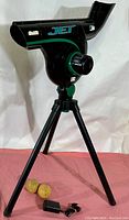 Front view of Atec Jet pitching machine on tripod with two beige baseball practice balls and power adapter on floor.