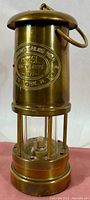 Full view of the upright brass lantern showing cylindrical shape, brass finish, hanging ring, and plaque on body.