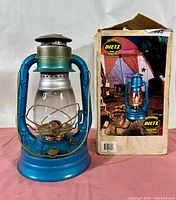 Front view of the blue Dietz No. 8 Air Pilot lantern next to its original cardboard box, showing details of the chimney, glass globe, wick, and base.