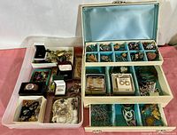 Clear plastic bin of assorted rings, bracelets, earrings on left; white jewelry box with blue velvet-lined trays on right