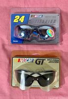 Two pairs of NASCAR branded sunglasses in sealed packaging on pink fabric background. Top pair is NASCAR GT, bottom pair is NASCAR Titanium with number 24 logo.