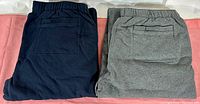 Two pairs of folded athletic sweat pants in navy blue and charcoal heather grey, viewed from back showing patch pockets and elastic waistbands.