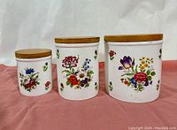 Three porcelain canisters of different sizes with floral country garden pattern and wooden lids arranged on a pink cloth.