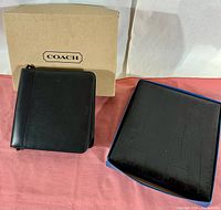 Coach black leather mini zipper portfolio with original box shown on a pink surface with white background.