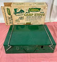 Green Coleman camp stove model 411A closed with original box in background, showing top and folded legs