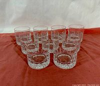 Front view showing all nine crystal whiskey tumblers arranged in a V shape on a reddish cloth background.