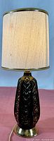 Full side view of the black and gold porcelain lamp with beige fabric shade on a pink surface with blue background.