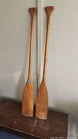 Full view of two wooden canoe paddles standing against a wall, showing overall length and shape.