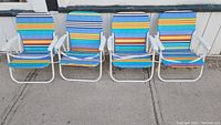 Four folding beach chairs with multicolor stripe design and white frame lined up on pavement.
