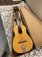 Front view of acoustic guitar in cardboard box on lace-patterned fabric. Wooden body with natural finish and metal tailpiece visible.