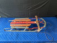 Full view of vintage Jetstream sled showing red and brown wooden slats over a metal red frame and metal runner base, lying on blue quilted surface.