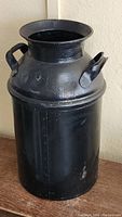 Full view of black painted metal milk can showing cylindrical shape, side handles, and flared opening.
