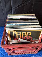 Photo showing multiple vinyl records stored upright inside a red plastic crate with visible wear and manufacturer markings.