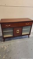Front view of the mid-century modern buffet showing one drawer with brass handles and glass cabinet doors below with a solid wood center panel.