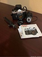 Photo showing the full lot contents displayed on a dark wood surface including camera, manual, case, tripod and light meter.