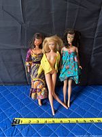 Three vintage dolls standing against a dark backdrop with blue textured surface; ruler shows approximately 11.5 inch height.