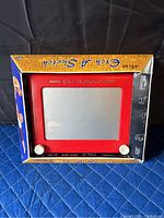 Front and angled view of vintage Magic Etch A Sketch toy in original packaging, showing red frame and white knobs.