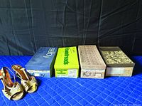 Four vintage women’s shoe boxes and one pair of beige leather slingback heels on blue quilted surface with black backdrop.