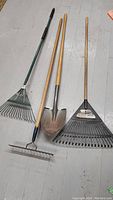 Full view of the three yard tools laid on the floor: green-handled metal leaf rake, metal garden rake with wooden handle, and square-point shovel with wooden handle.