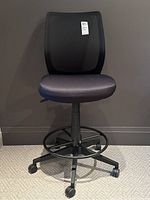 Front view of black rolling drafting chair showing mesh backrest, padded fabric seat, circular foot ring, and caster wheel base.