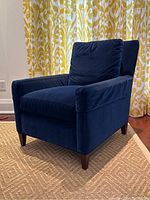 Three-quarter front view of royal blue velvet lounge chair on woven rug with patterned curtains behind