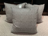 Three grey and white geometric patterned square pillows arranged in front of a wooden shelf showing their down fluffy fill and zippered covers.