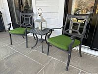 Two black cast aluminum chairs with green cushions and a matching lattice side table holding a stainless steel lantern