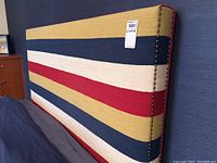 Close-up angled view showing bold horizontal stripes in mustard yellow, navy blue, red, and cream upholstery and brass-tone nailhead trim on side edge