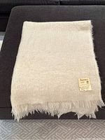 Cream mohair throw blanket folded and resting on a dark fabric ottoman, showing a fabric label on one corner.