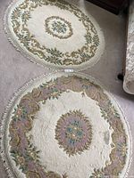 Photo showing both circular floral area rugs placed on carpeted floor with fringe edges, predominantly cream background with pink, green and blue floral patterns.