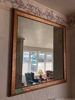 Full view of large square framed mirror reflecting a window and furniture with a gold detailed wooden frame.