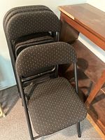 Four black folding chairs stacked together. Chairs have black fabric upholstery with small white dot pattern and black metal frames.
