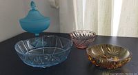 Four colored glass pieces arranged on a black surface: aqua pedestal covered dish, translucent pink swirl bowl, blue footed bowl, amber cut glass candy dish.