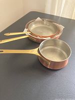 Three copper pans with brass handles displayed on a flat surface, showing interiors and exteriors