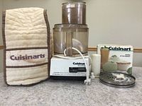 Photo showing all items in lot: quilted cover labeled 'Cuisinarts', main food processor base with clear plastic bowl, whisk attachment box, and white plastic attachments on countertop.