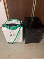 Two dehumidifiers placed side by side, black Mastercraft D38 Frostguard and white Classic 70 pint model with green cord.